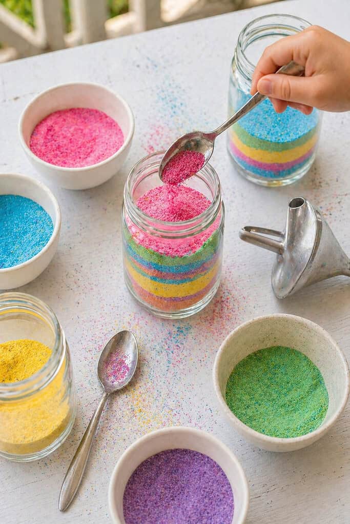 a small clear glass jar being filled with layered colored sand