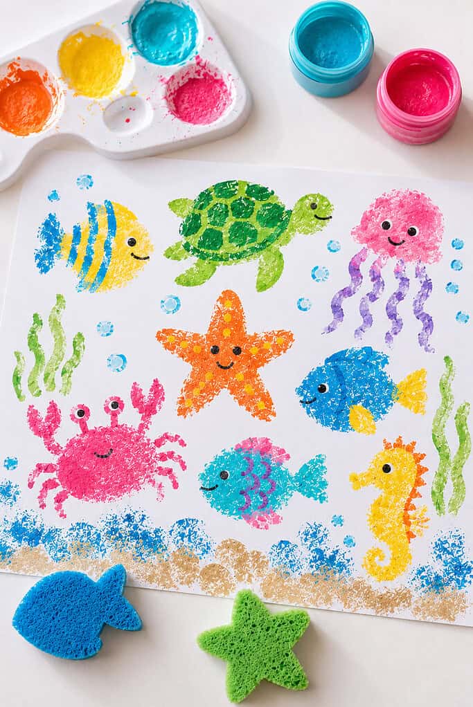 A large sheet of white paper filled with sponge-painted sea creatures such as fish, starfish, crabs, turtles, and jellyfish created using cut kitchen sponges dipped in bright paint.