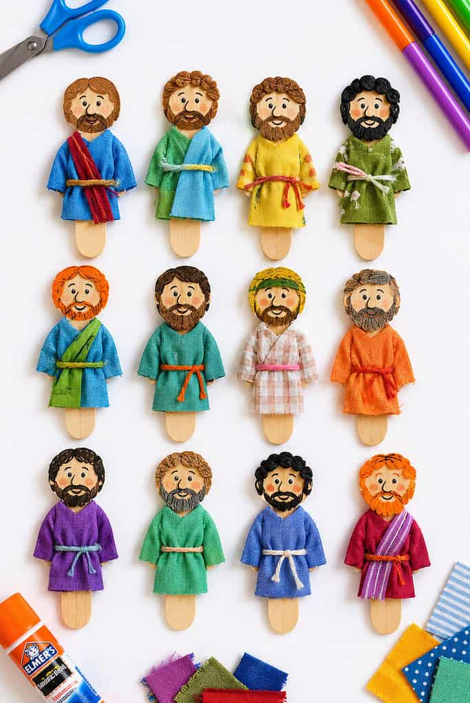 a wooden popsicle stick and represent one of the Twelve Disciples, with a unique hand-drawn or printed face, simple hair and beard details, and a small colorful fabric robe made from scrap cloth.