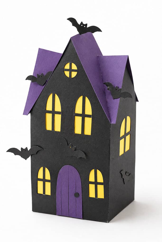 A 3D black paper house box with a purple roof, yellow arched windows, a purple arched front door, a circular window at the top, and small black paper bat cutouts flying around the exterior