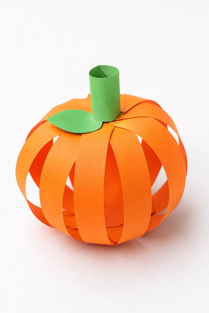A 3D round shape made from curved orange paper strips fanned out in a sphere, topped with a green paper tube stem and a small green paper leaf cutout