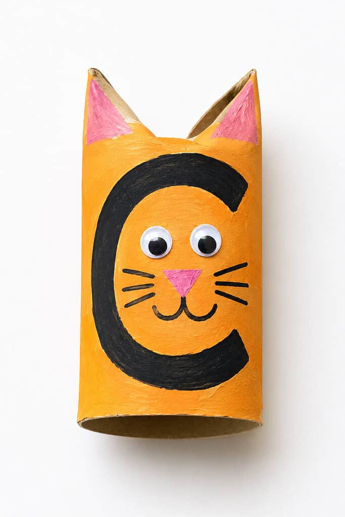 Orange toilet paper roll painted with a large black letter C, two pointy ears pinched at the top with pink inner ear details, two googly eyes, drawn whiskers, and a pink triangle nose
