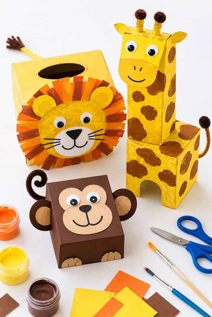 a small collection of handmade zoo animals created from recycled cardboard boxes, including a tissue box lion with a fluffy paper mane, a cereal box giraffe with painted spots and a long neck, and a small jewelry box monkey with playful facial features