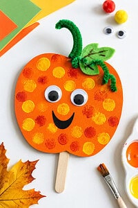 A round orange paper plate craft with finger-painted orange dots, two large googly eyes, a black curved smile, and a green pipe cleaner stem, resting on a white surface alongside autumn leaves, a paintbrush, and an open orange paint container.