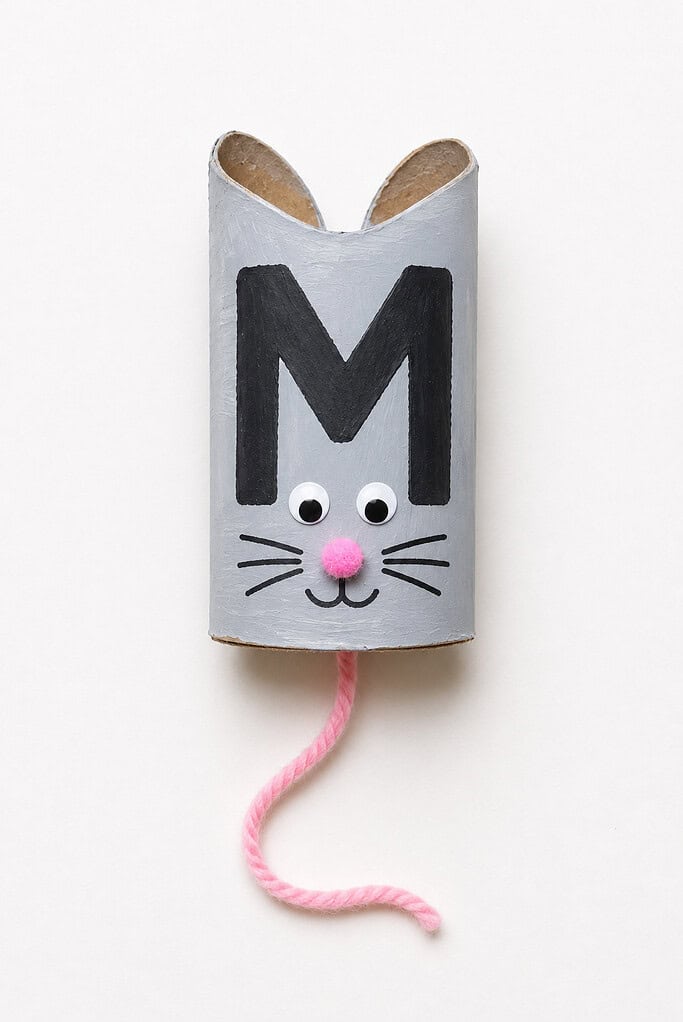 Gray painted toilet paper roll standing upright with two pointed ears pinched at the top showing brown inner ear color, a bold black letter M painted on the front face, two googly eyes, a pink pom-pom nose, drawn whiskers, and a long pink yarn string tail hanging from the bottom