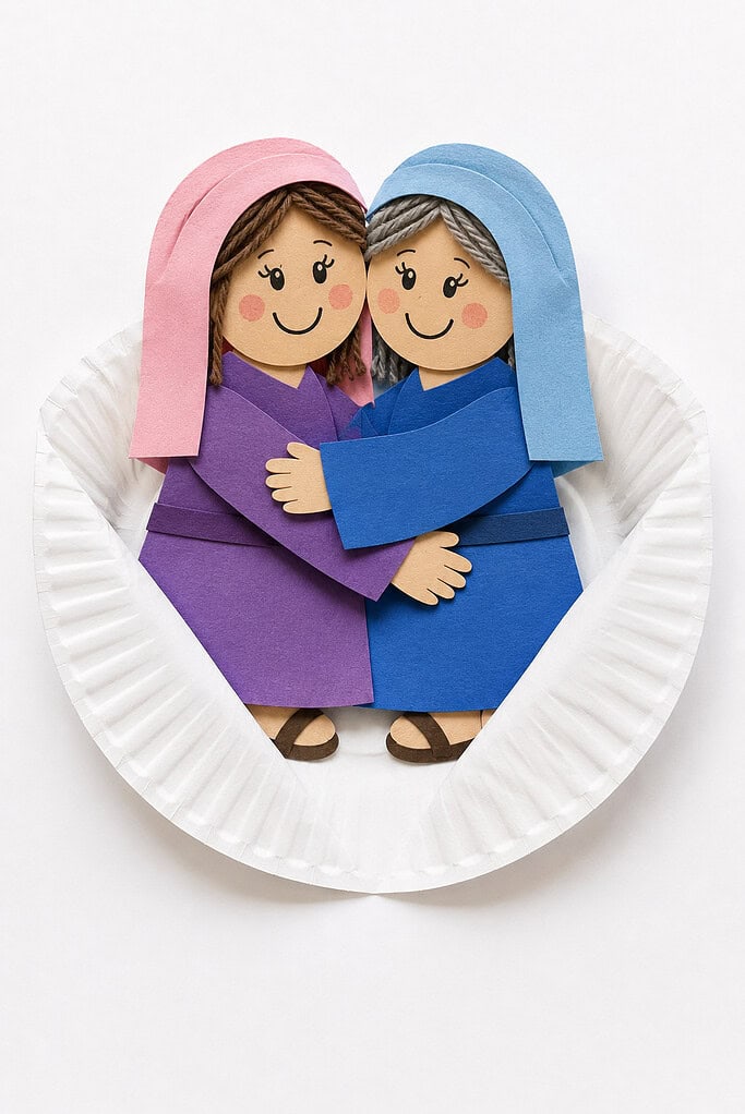 Two paper cutout female figures hugging each other on a white paper plate; the left figure wears a purple robe and pink headscarf with brown hair, and the right figure wears a blue robe and blue headscarf with gray hair, both with drawn smiling faces and brown sandals