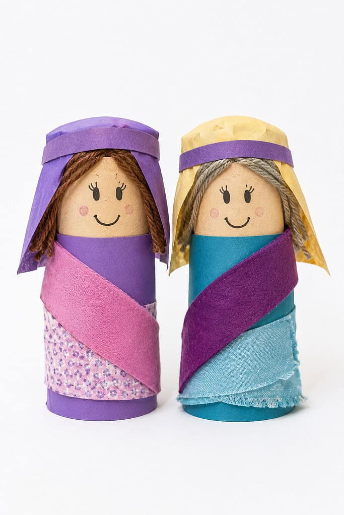 Two toilet paper roll puppets decorated as female figures; the left one is wrapped in purple paper with brown yarn hair and a purple headscarf, wearing a pink sash and floral fabric skirt; the right one is wrapped in teal fabric with gray yarn hair and a yellow headscarf with purple band, wearing a purple sash