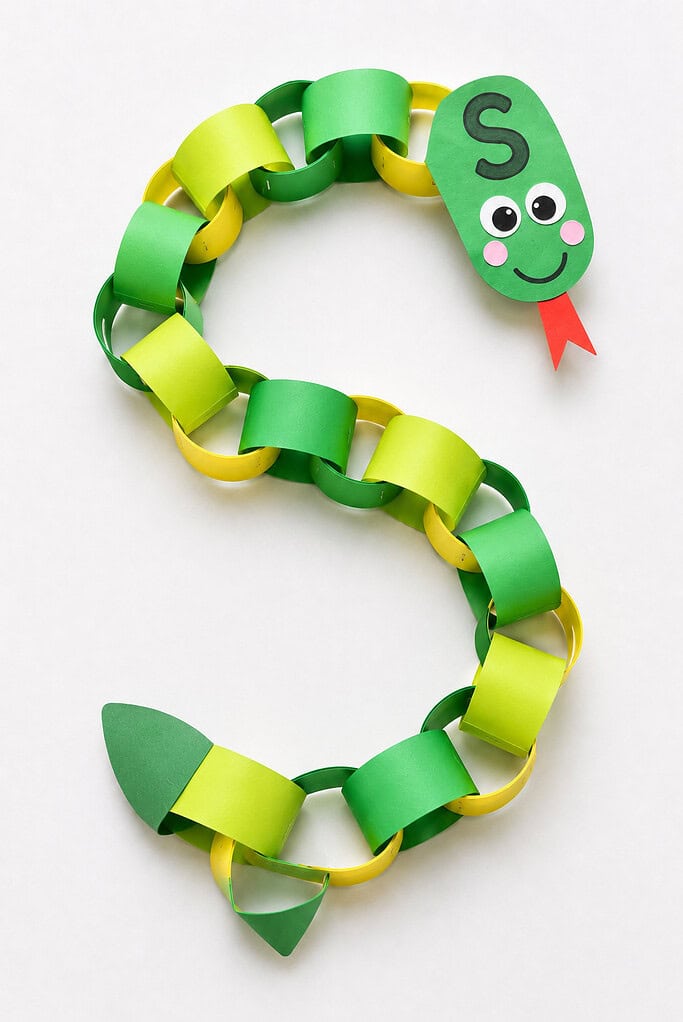 Green and yellow paper chain links looped into a long S-curve with a dark green oval head at the upper right showing two googly eyes, pink cheeks, a smiling mouth, and a red forked paper tongue, and a pointed green paper tail at the lower left end
