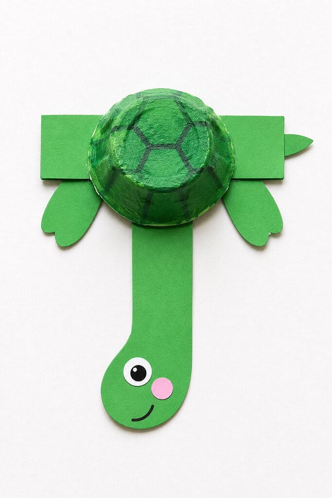 Green paper letter T with a dome-shaped painted egg carton cup placed on the crossbar as a shell with dark green hexagonal markings, two green scalloped flipper shapes on each side of the crossbar, a small green rounded head at the bottom of the vertical stroke with a single googly eye, a pink cheek circle, and a small curved smile
