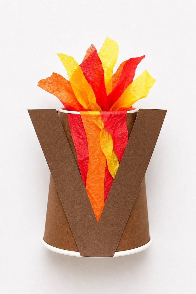 Brown paper letter V shape attached to the rim of a white paper cup with the V's two angled sides forming the mountain walls, and crinkled tissue paper in red, orange, and yellow scrunched and erupting out of the top of the cup