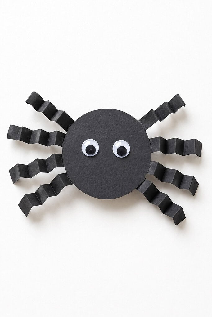 A black circular paper body with eight accordion-folded black paper legs extending outward on all sides and two small googly eyes in the center