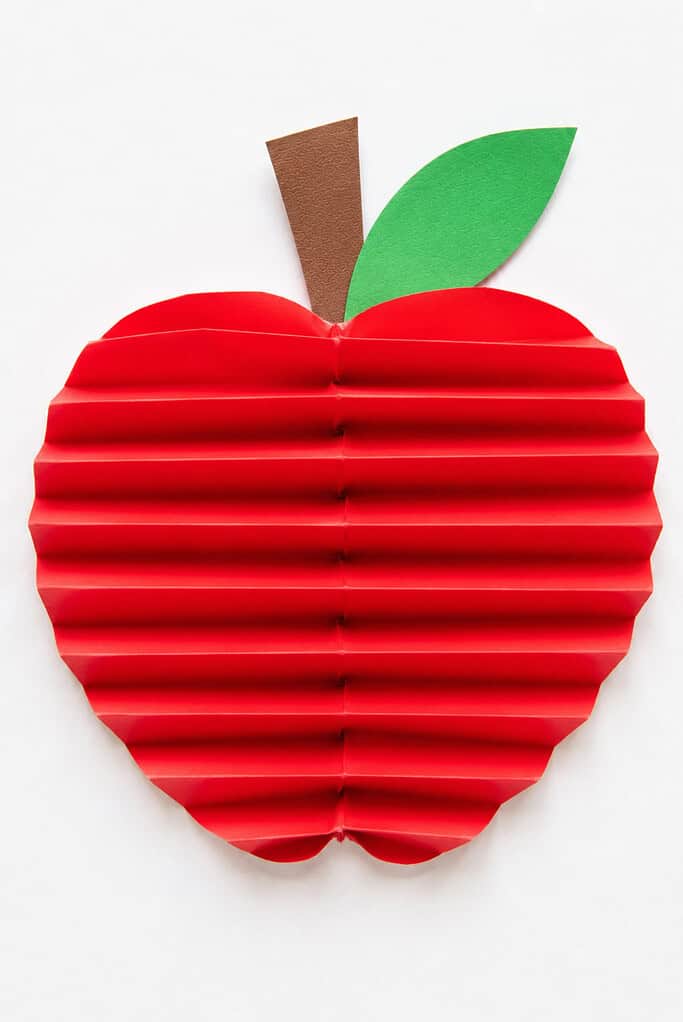 An apple-shaped red paper with horizontal accordion folds creating a ribbed texture, topped with a brown paper stem and green paper leaf