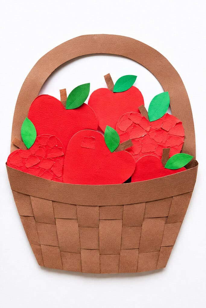 A brown woven paper basket with a looped handle, filled with multiple red paper apple shapes with brown stems and green leaves