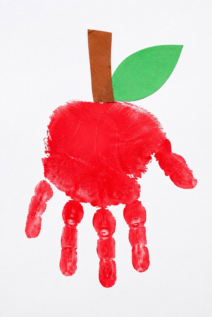 A red handprint on white paper with fingers pointing downward, topped with a brown paper stem and green paper leaf