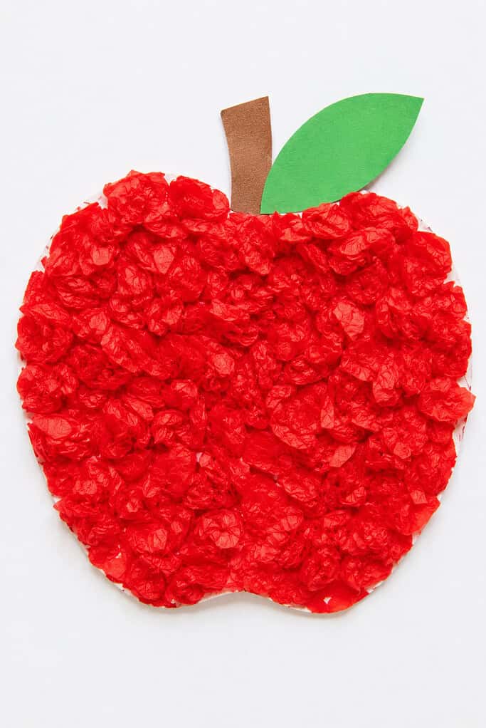 An apple shape filled with crumpled red tissue paper pieces glued over the surface, with a brown paper stem and green paper leaf on top