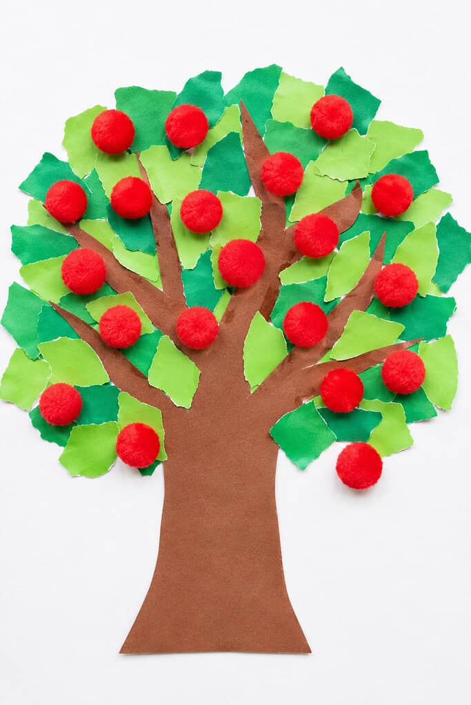 A paper tree with a brown trunk and branches, covered with torn green paper pieces for leaves and red pom-poms scattered throughout the canopy