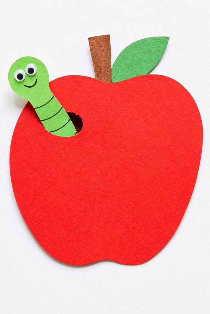 A large red foam apple cutout with a brown stem and green leaf, featuring a small hole with a green foam worm with googly eyes and a drawn smile poking out of it