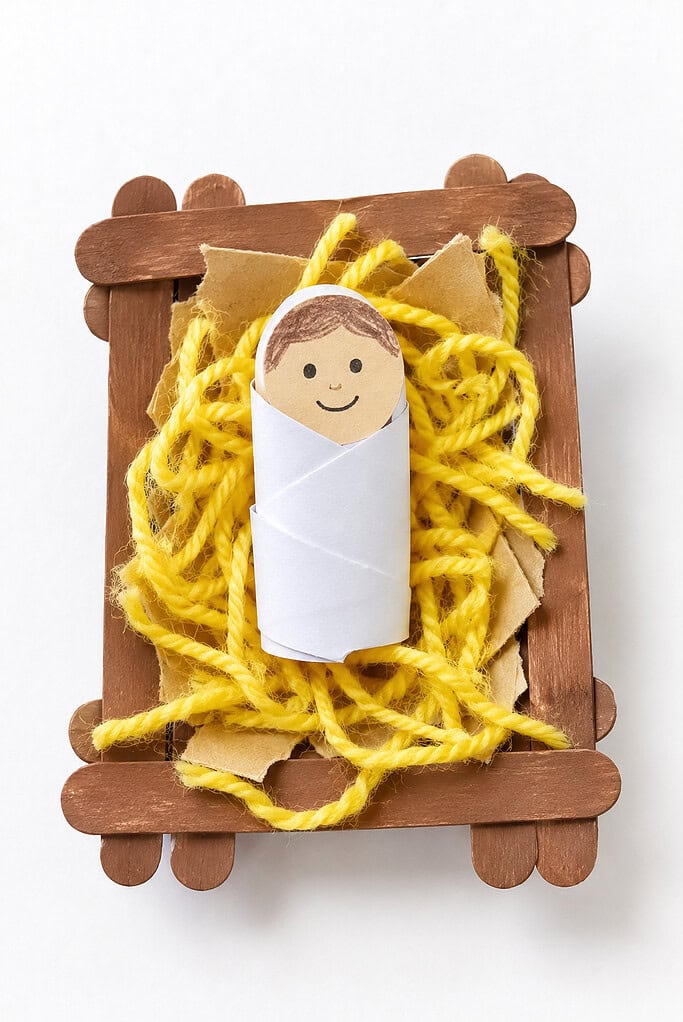 a tiny handmade manger constructed from a few wooden craft sticks or a small brown cardboard box, filled with bright yellow yarn strands and torn paper pieces resembling soft hay.