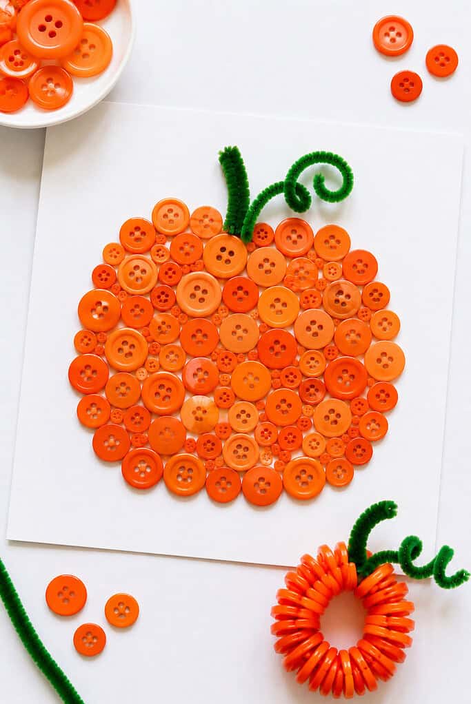 A handmade pumpkin craft created from an assortment of vibrant orange buttons arranged into a classic pumpkin shape on a square piece of white cardstock.