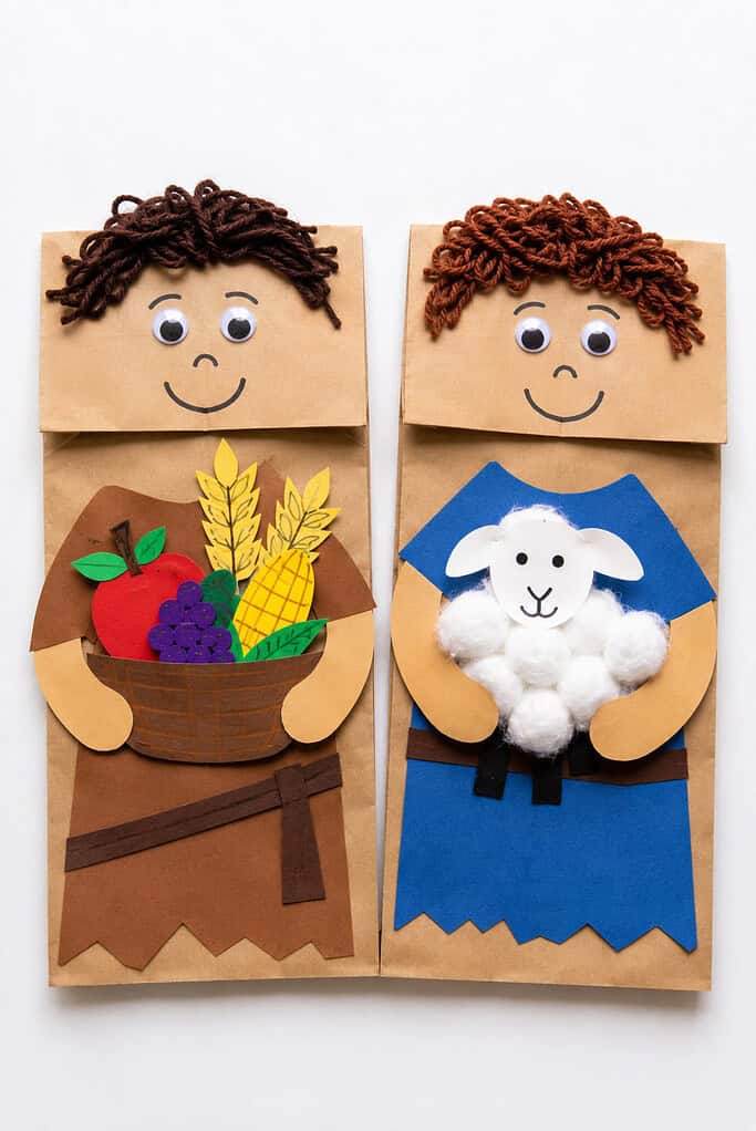 Two paper bag puppets placed side by side. Each puppet has a round face with yarn hair, googly eyes, and a drawn smile. The left puppet wears a brown paper outfit and holds a paper basket filled with cut-out fruits and grain. The right puppet wears a blue paper outfit and holds a small white cotton ball lamb.