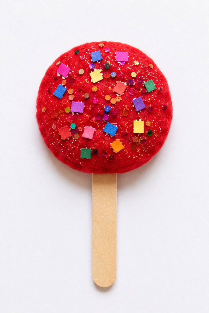A round red felt circle covered in gold glitter and colorful sequins, attached to a wooden craft stick