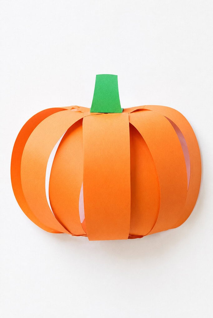 A 3D pumpkin shape made from arched orange construction paper strips fanned out in a round ball shape, with a small green paper cylinder at the top as a stem