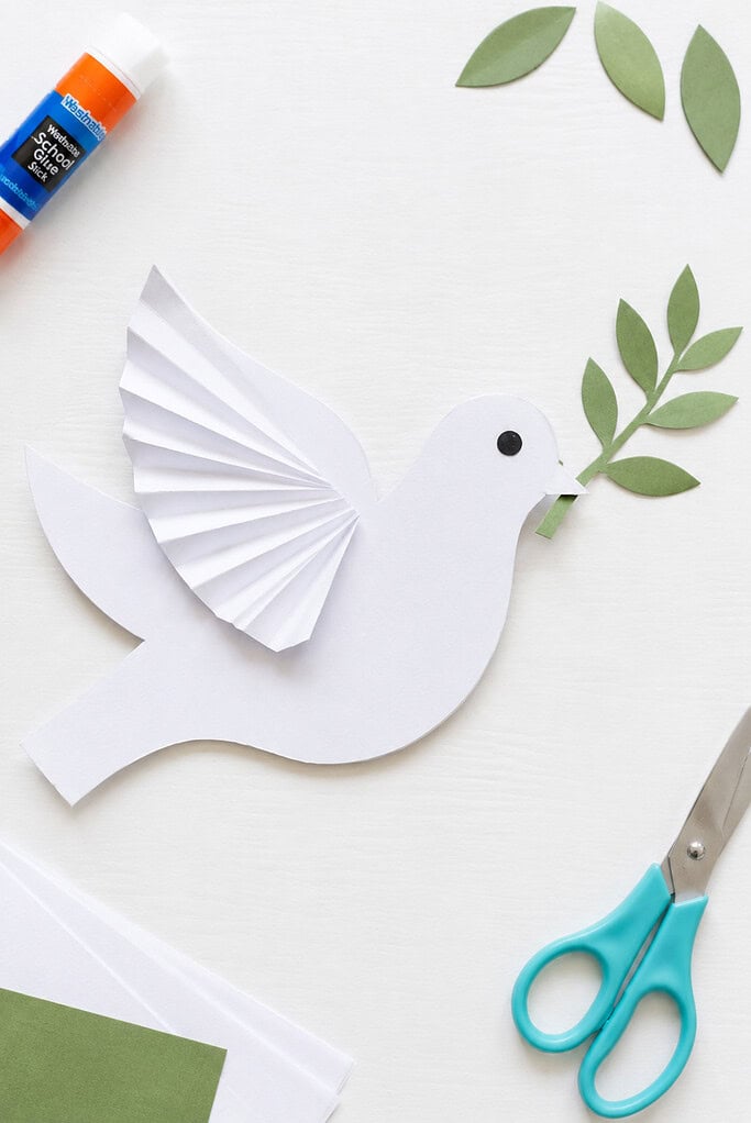 a simple handmade white paper dove created by a child, featuring a folded-paper wing for dimension and a small green olive branch held gently in its beak.