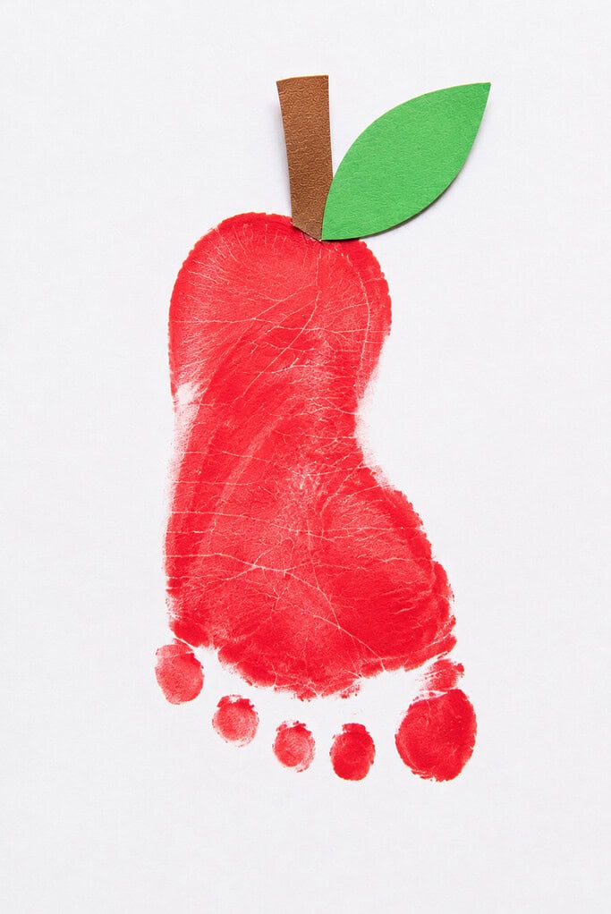 A red footprint painted on white paper with toes pointing downward, topped with a brown paper stem and green paper leaf