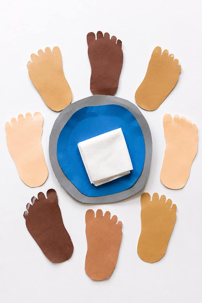 a simple paper basin made from hand-cut blue and gray construction paper, a small paper towel crafted from folded white paper, and several child-traced paper feet cut from construction paper.