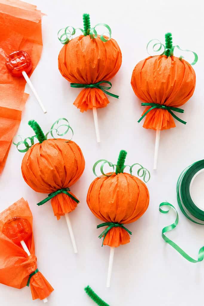 Several round lollipops wrapped in vibrant orange tissue paper and tied at the base with green ribbon, twine, or pipe cleaners to create miniature pumpkin-shaped treats.