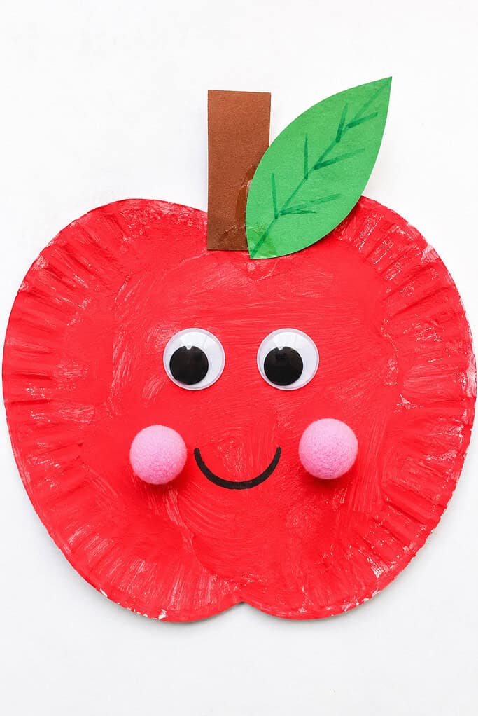 A red-painted paper plate shaped like an apple face, with googly eyes, pink pom-pom cheeks, a drawn smile, a brown paper stem, and a green paper leaf on top