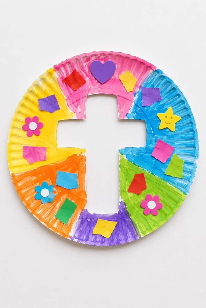 A round paper plate painted in multicolored sections with a cross-shaped cutout in the center, decorated with foam stickers including hearts, flowers, and stars