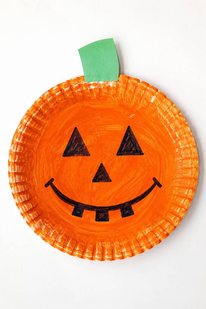 A paper plate painted orange with triangular eyes, a triangular nose, and a smiling mouth with square teeth drawn in black marker, with a green paper stem attached at the top