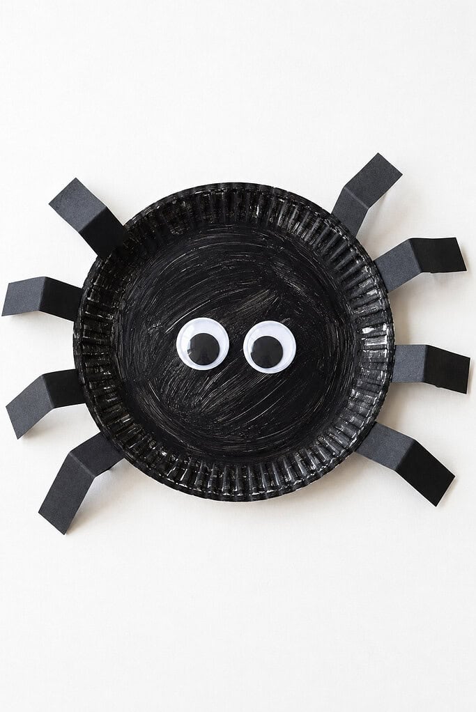 A paper plate painted black with eight bent black paper legs attached around the sides and two large googly eyes in the center