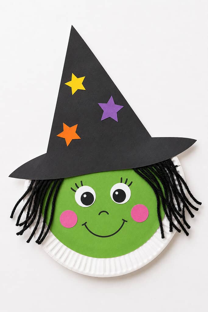 A paper plate with a green oval face with googly eyes with drawn eyelashes, pink circular cheeks, and a smile, with black yarn hair hanging on the sides and a tall black paper pointed hat decorated with yellow, orange, and purple paper stars