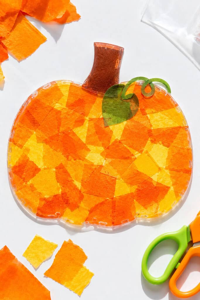 A handmade pumpkin suncatcher created from torn pieces of vibrant orange and golden yellow tissue paper sealed between two layers of clear contact paper cut into a pumpkin shape.