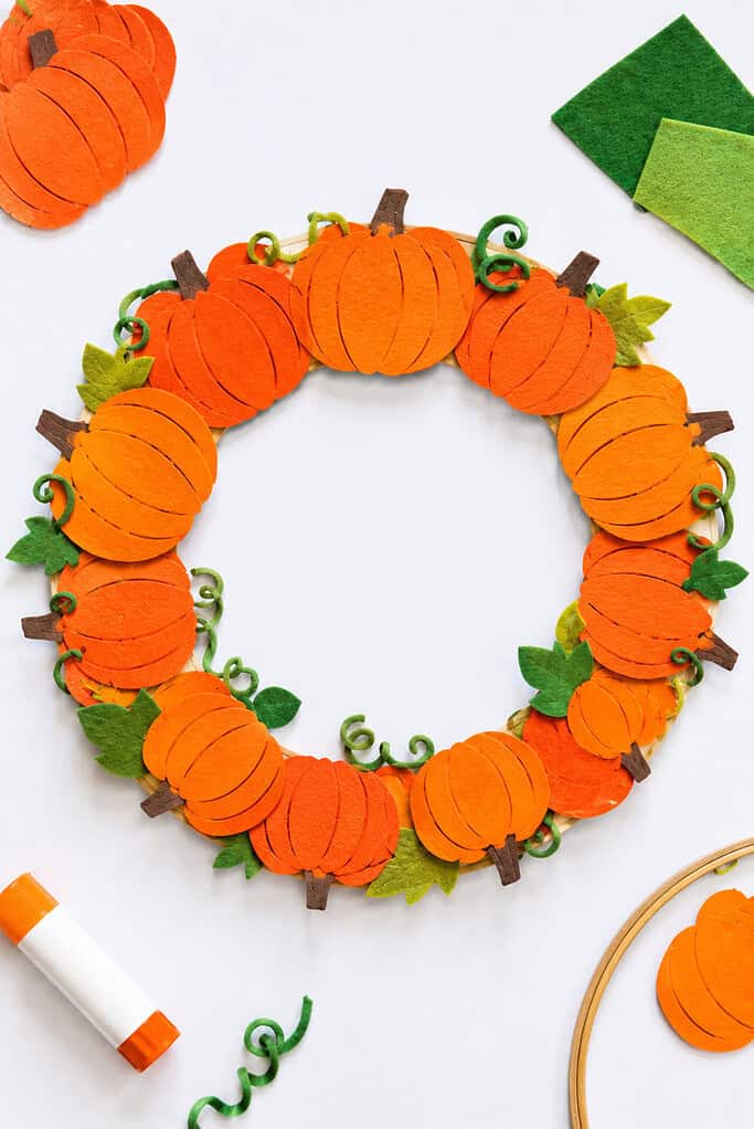 A handmade autumn wreath created by attaching multiple pumpkin cutouts made from vibrant orange paper or soft felt around an embroidery hoop or cardboard ring.
