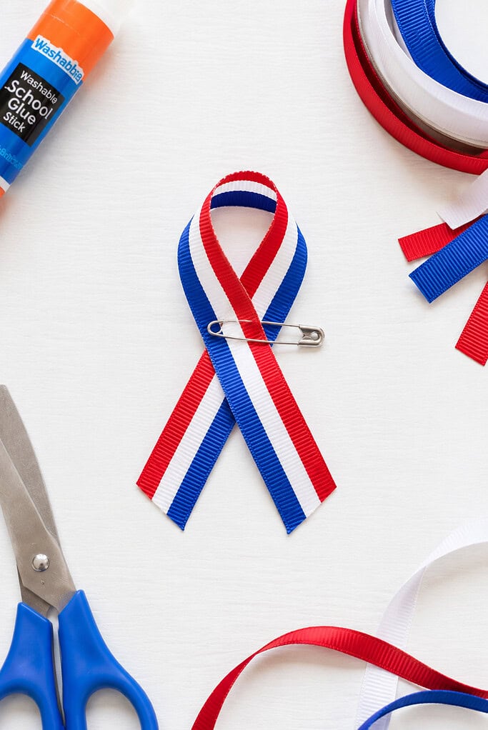 a simple handmade awareness-style ribbon pin crafted from red, white, and blue ribbon, neatly folded into a loop and secured with a small safety pin or a discreet dab of glue.