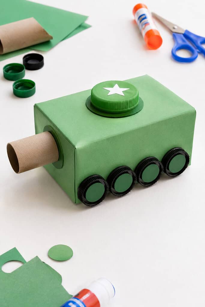 a playful tank made from recycled materials, including an empty tissue box wrapped in green construction paper, a cardboard tube cannon, and bottle-cap wheels.