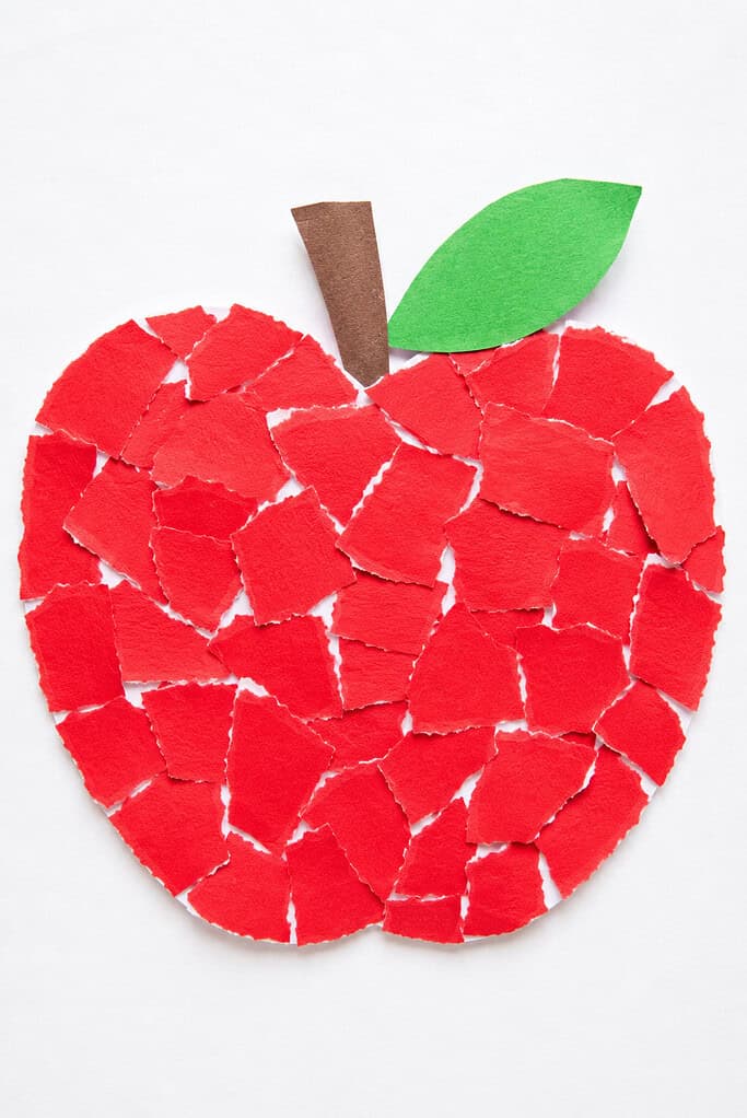 An apple shape filled with torn pieces of red paper arranged like a mosaic, with a brown paper stem and a green paper leaf on top