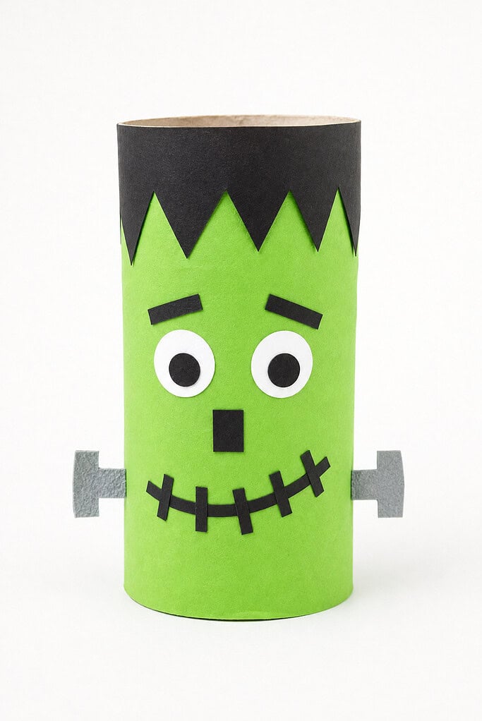 A toilet paper roll wrapped in green paper with a black zigzag hair strip at the top, white circular eyes with black pupils, black rectangular eyebrows, a black square nose, a stitched smile, and gray paper bolts on both sides
