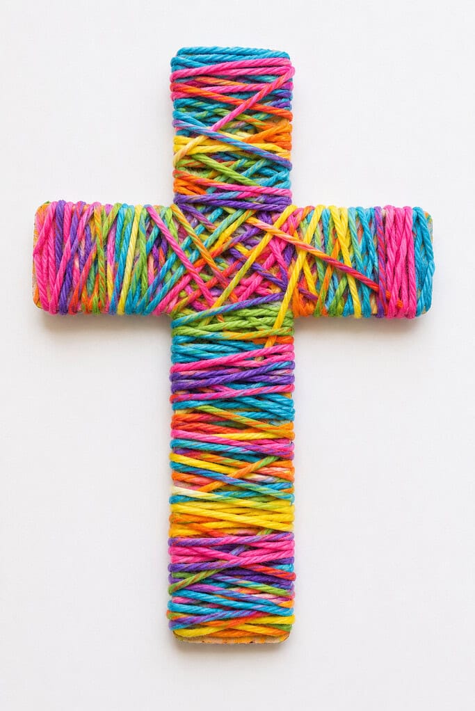 A wooden cross shape fully wrapped with multicolored yarn in pink, teal, yellow, purple, green, and orange