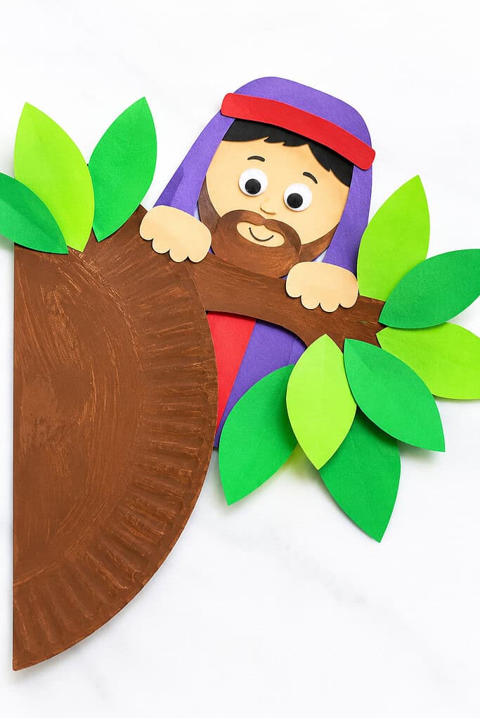 A paper craft featuring a smiling bearded figure with a purple robe and red headband, peeking over a brown painted paper plate tree trunk, surrounded by green paper leaves