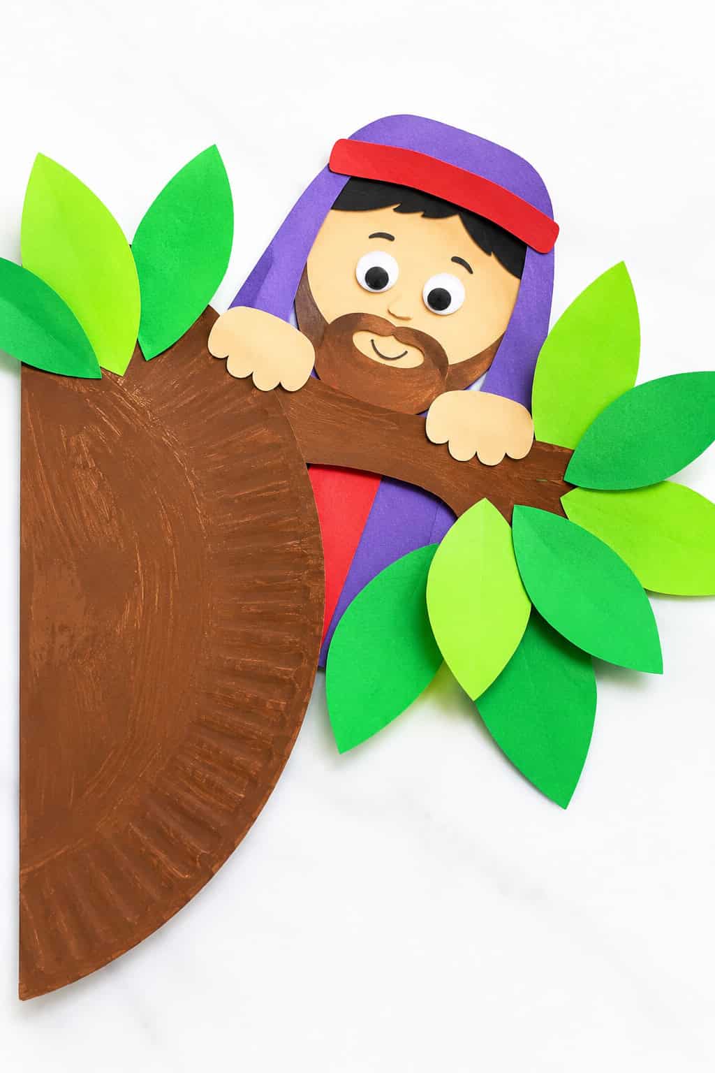 A paper craft featuring a smiling bearded figure with a purple robe and red headband, peeking over a brown painted paper plate tree trunk, surrounded by green paper leaves