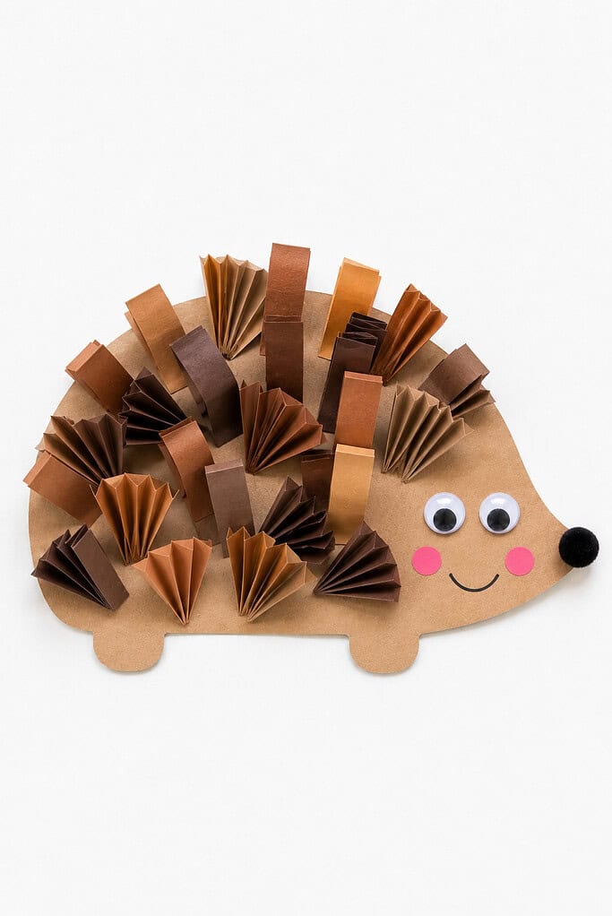 Accordion-folded paper fans in shades of brown arranged as 3D spikes on a tan paper hedgehog body, with googly eyes, a black pom-pom nose, and pink cheeks