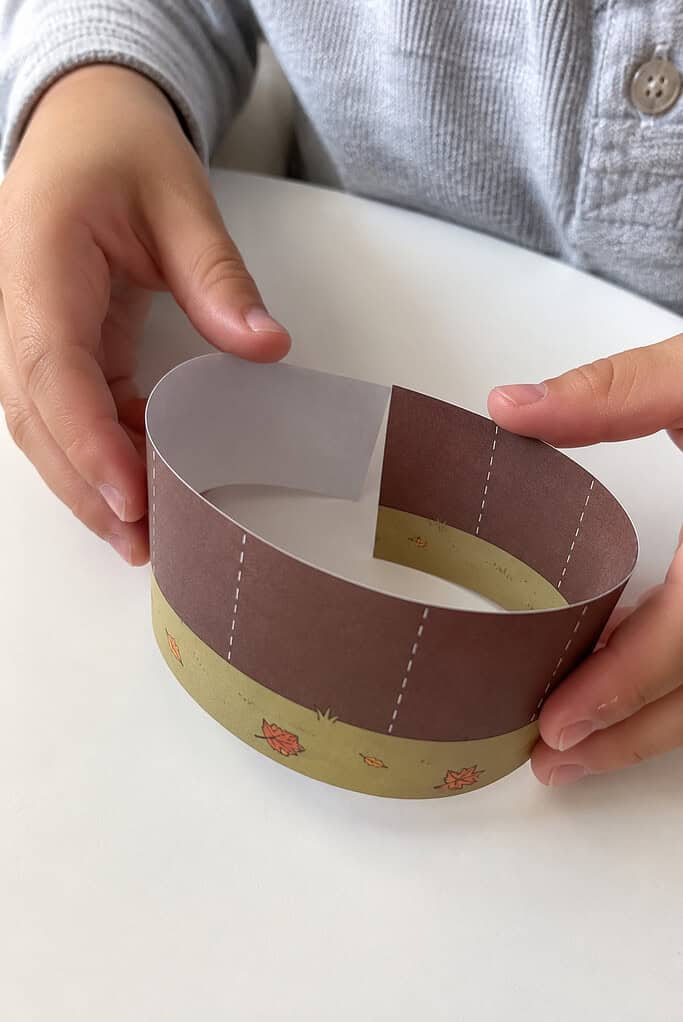 A child's hands folding a printed paper strip with a brown top and green grass bottom into a ring on a wooden table