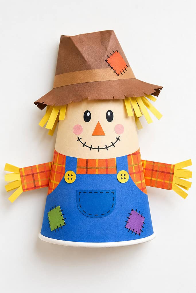 Paper cup figure on a white background with a tan face, yellow paper-strip hair, black oval eyes, an orange triangular nose, pink cheeks, and a stitched smile. It wears a brown paper hat with a tan band and orange patch, an orange plaid shirt, and blue overalls with yellow buttons, a front pocket, green and purple patches, and outstretched arms with yellow paper-strip ends.