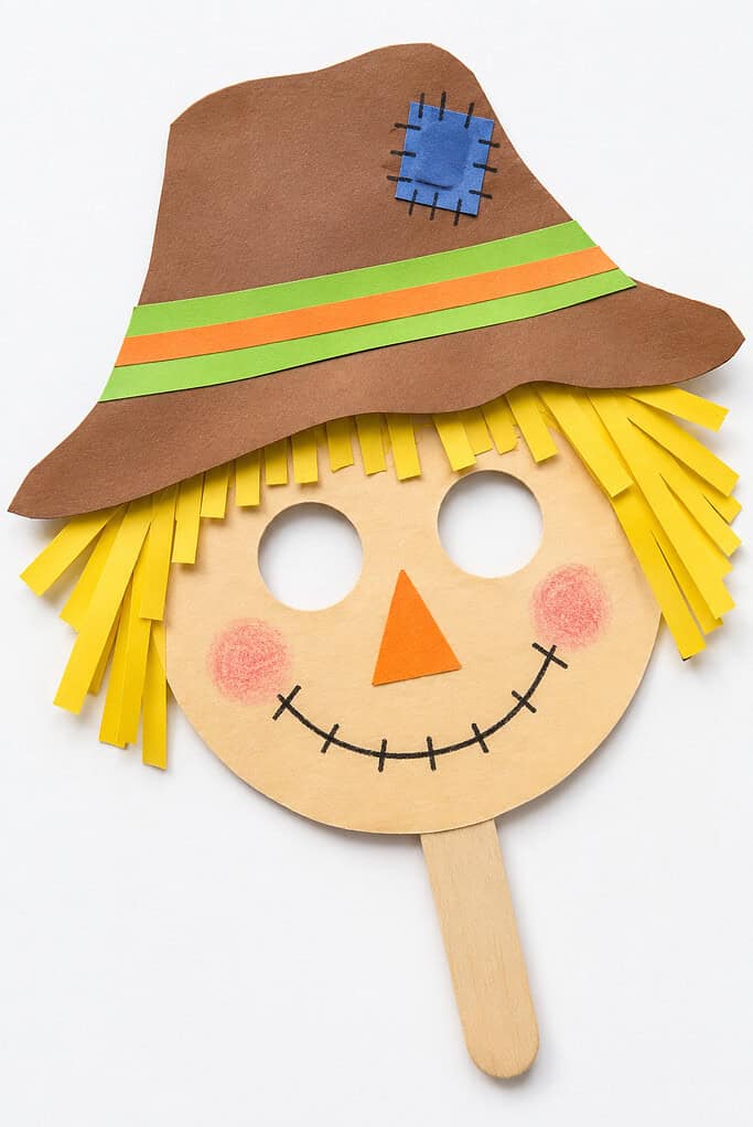 Paper face cutout on a white background with two circular eye holes, yellow paper-strip hair, an orange triangular nose, pink cheeks, and a stitched smile. It wears a large brown hat with green and orange bands and a blue stitched patch, with a wooden craft-stick handle extending below.