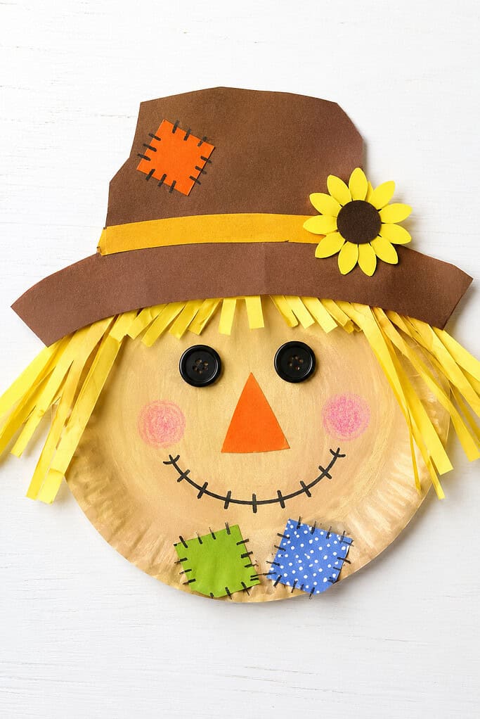 Paper plate face with yellow paper-strip hair, black button eyes, an orange triangular nose, pink cheeks, a stitched smile, and green and blue patches. A large brown paper hat with a yellow band, orange patch, and yellow flower sits above the face on a white wooden surface.