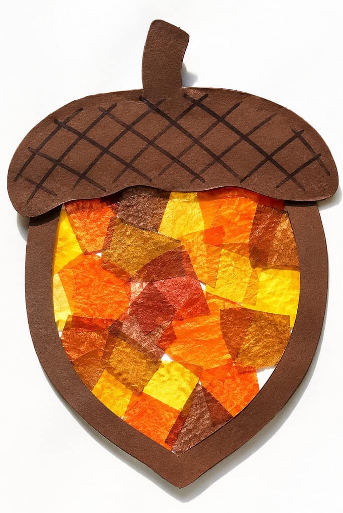 A layered paper acorn with a dark brown cap and border, a short stem, and black crisscross lines across the cap. The center is filled with overlapping translucent tissue-paper squares in yellow, orange, red, and brown on a white background.